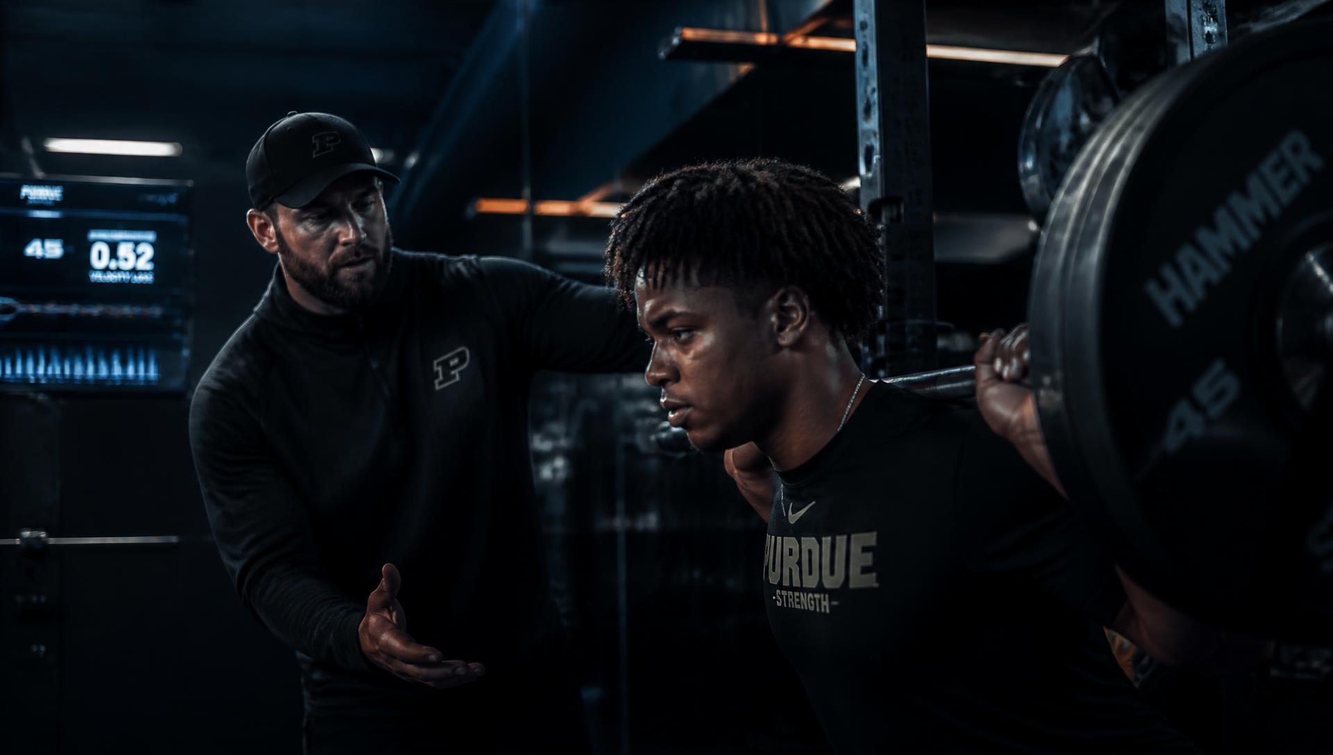 Strength coach guiding an athlete through a barbell set in a dark performance gym